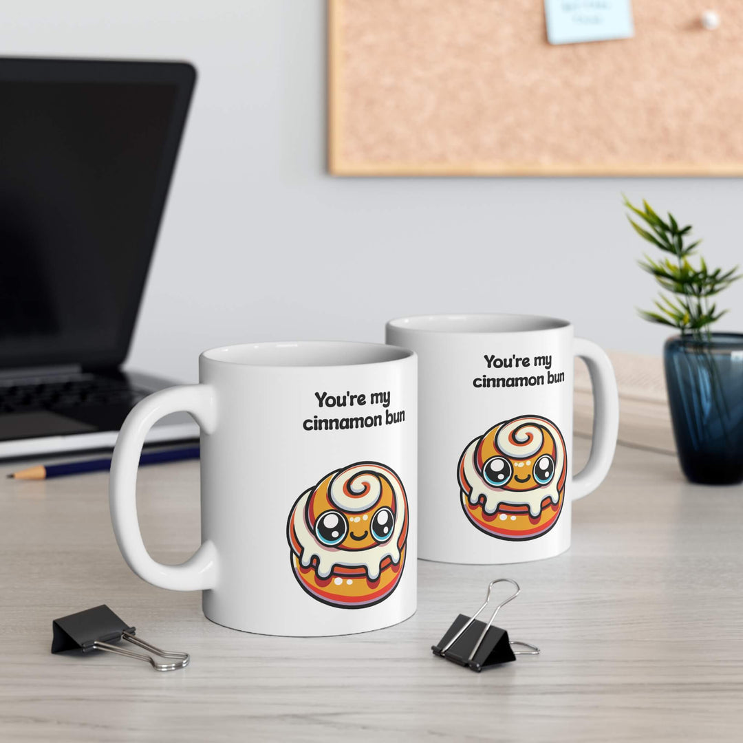 A charming ceramic mug with the phrase "You're my cinnamon bun" and a kawaii-style cinnamon bun illustration, set in a cozy holiday setting with cinnamon sticks and pine cones, enhancing a warm, festive atmosphere.

