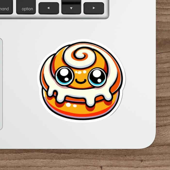 A kawaii-style doughnut sticker with vibrant orange glaze and swirling white icing, placed on a laptop next to a keyboard, embodying a cute and playful aesthetic