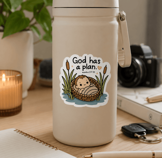 A beige insulated water bottle with a carry loop sits on a wooden desk in a styled room setting. The bottle features a die-cut sticker with a cute cartoon baby in a reed basket and the text &
