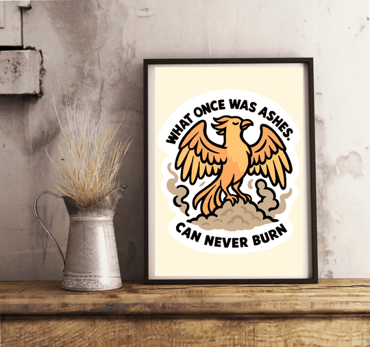 Framed wall art print featuring a bold orange phoenix rising from ashes, with the quote "What once was ashes can never burn" in strong black lettering, set against a light neutral background.