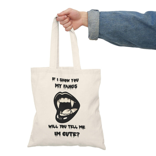A natural canvas tote bag showcasing a cheeky design with a cartoonish mouth displaying fangs and the text 'If I show you my fangs, will you tell me I'm cute?' The playful combination of humor and confidence makes this tote a great conversation starter."