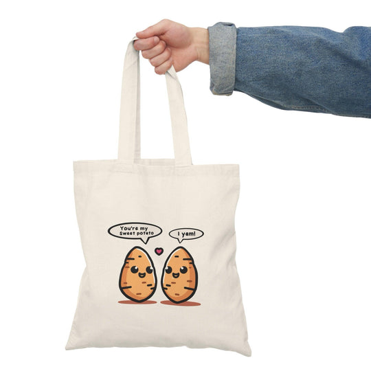 A cream-colored tote bag featuring a whimsical illustration of two sweet potatoes with smiling faces. The cute design adds a playful touch to this bag, appealing to food enthusiasts and anyone who enjoys a bit of fun in their accessories