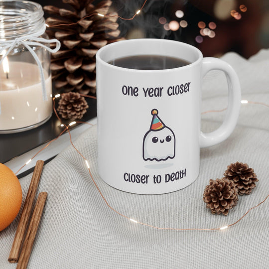 White ceramic mug featuring a ghost illustration and the darkly humorous text 'One Year Closer to Death,' perfect for those who enjoy a bit of morbid humor