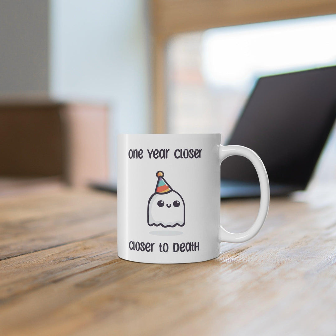 White ceramic mug featuring a ghost illustration and the darkly humorous text 'One Year Closer to Death,' perfect for those who enjoy a bit of morbid humor