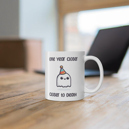 White ceramic mug featuring a ghost illustration and the darkly humorous text 'One Year Closer to Death,' perfect for those who enjoy a bit of morbid humor