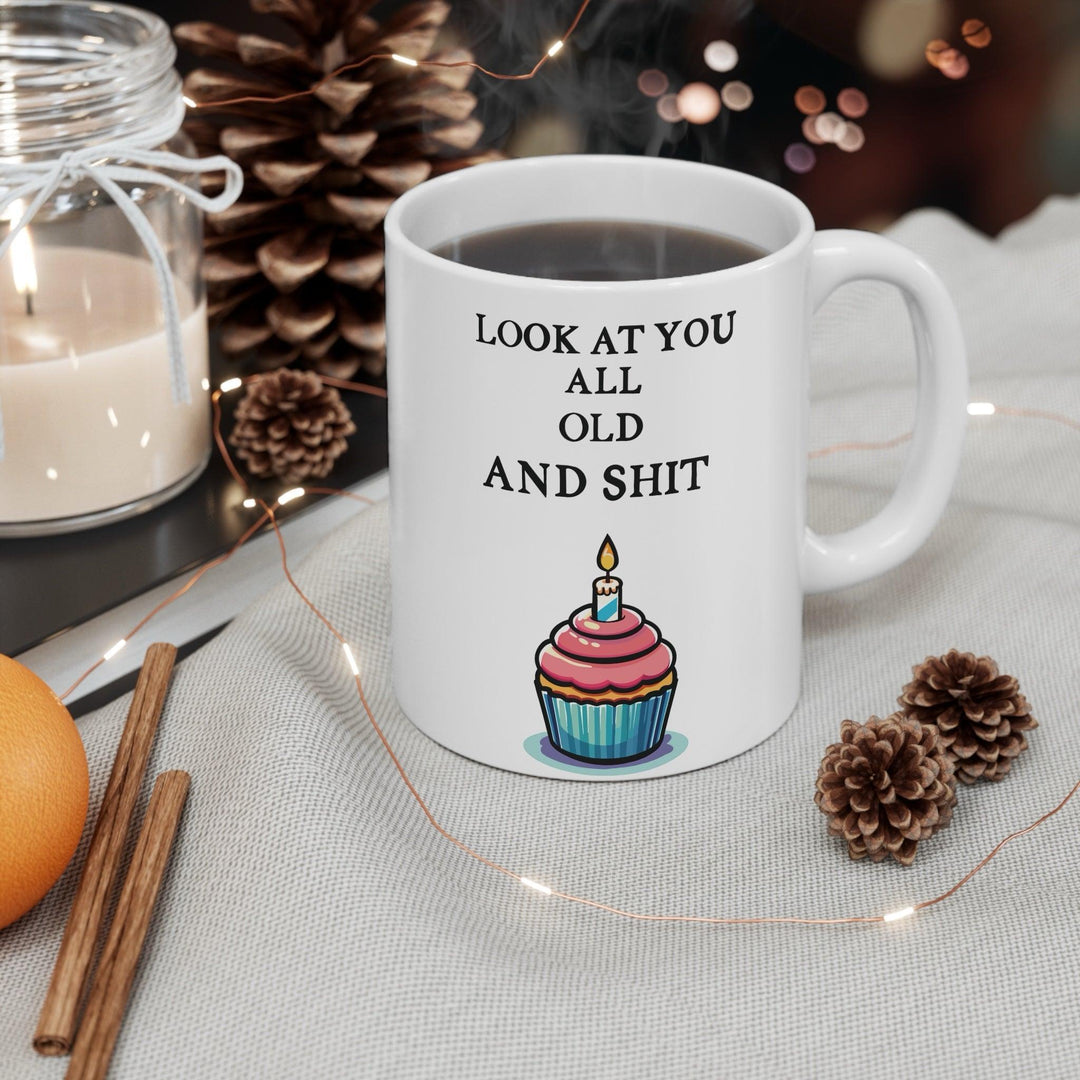 White ceramic mug featuring a playful cupcake design and the humorous phrase 'Look at You All Old and Shit,' perfect for adding some fun to a birthday celebration.