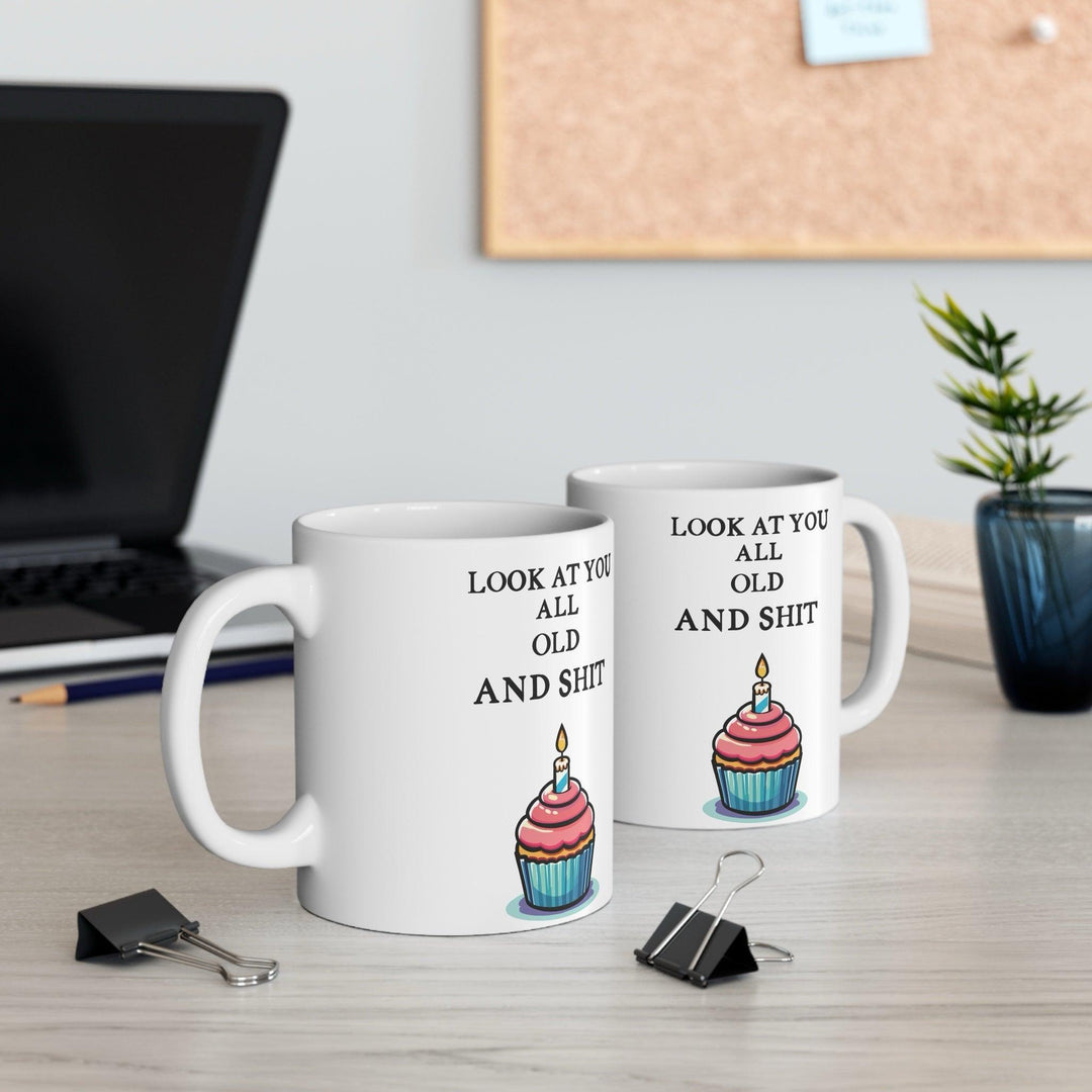 White ceramic mug featuring a playful cupcake design and the humorous phrase 'Look at You All Old and Shit,' perfect for adding some fun to a birthday celebration.