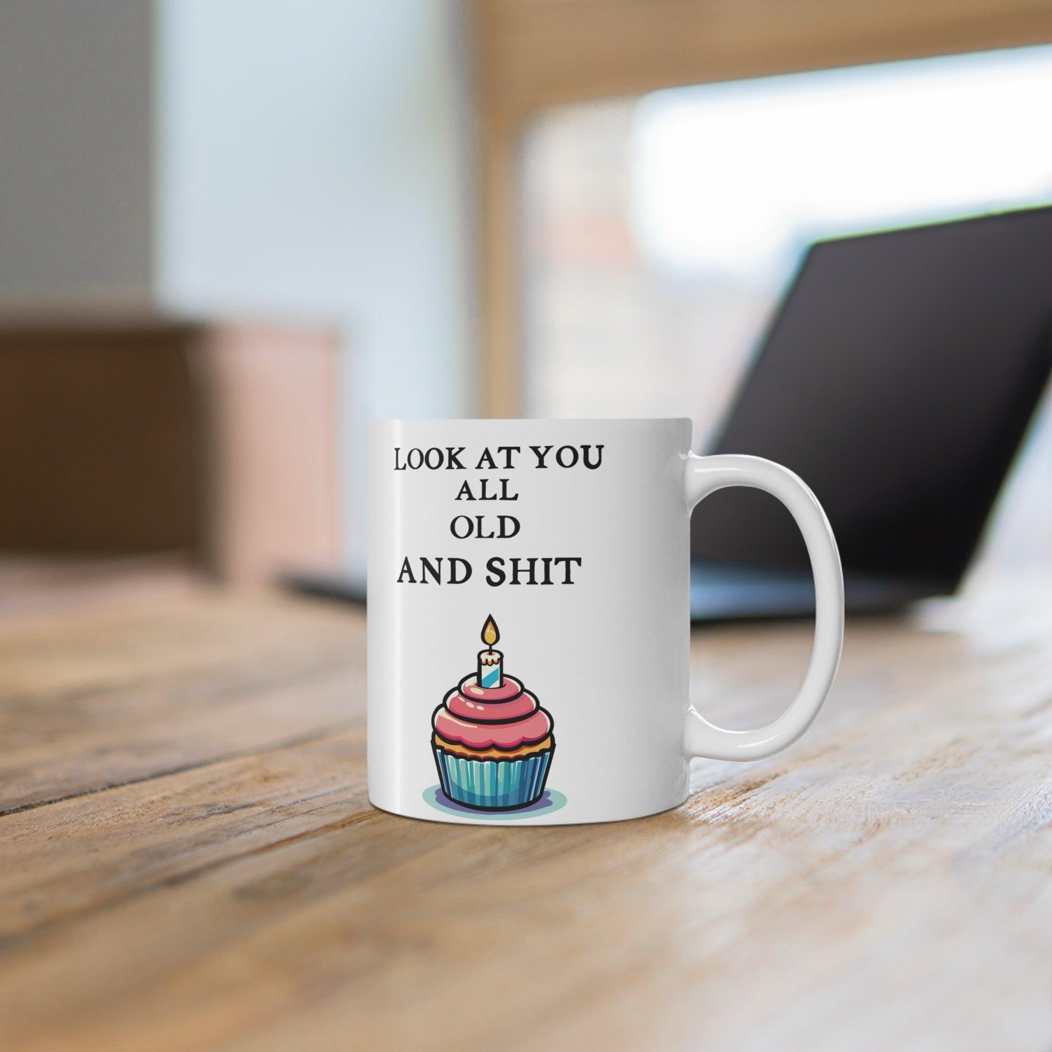 White ceramic mug featuring a playful cupcake design and the humorous phrase 'Look at You All Old and Shit,' perfect for adding some fun to a birthday celebration.