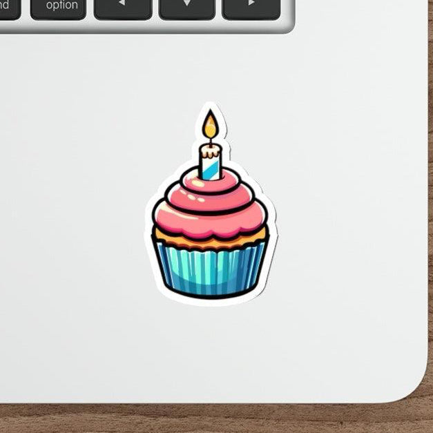 A sticker depicting a cartoon cupcake with a candle on top, placed on a laptop next to a mouse, representing the product's use as a decorative item for personalizing laptops and other surfaces.