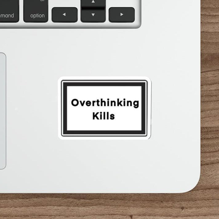 A sticker with the text 'Overthinking Kills' placed on a laptop next to a keyboard and a mouse, on a wooden desk.