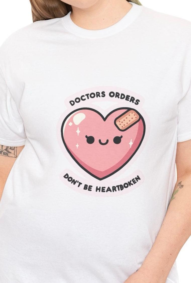 White T-shirt featuring a cute pink heart character with a bandage, accompanied by the phrase 'Doctor's Orders: Don't Be Heartbroken.' A light-hearted design offering an encouraging message of emotional healing.