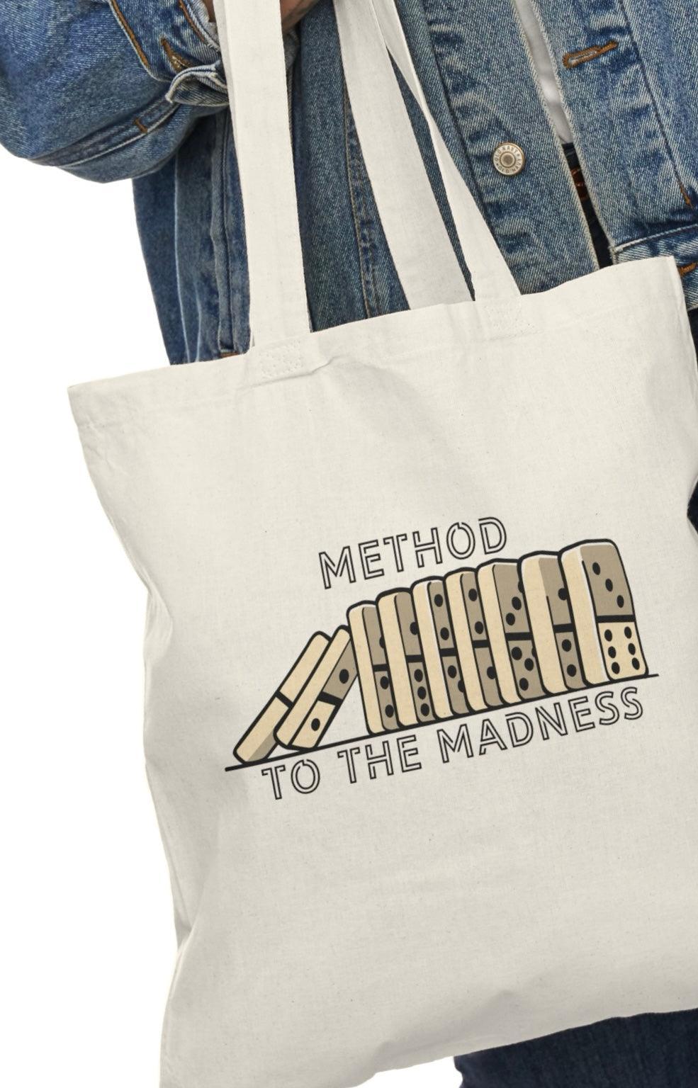 A cream-colored tote bag featuring a quirky design with the phrase 'Method to the Madness' accompanied by a whimsical illustration of a pencil and a notepad. The playful font and graphic emphasize a humorous take on creativity and the chaotic process of brainstorming