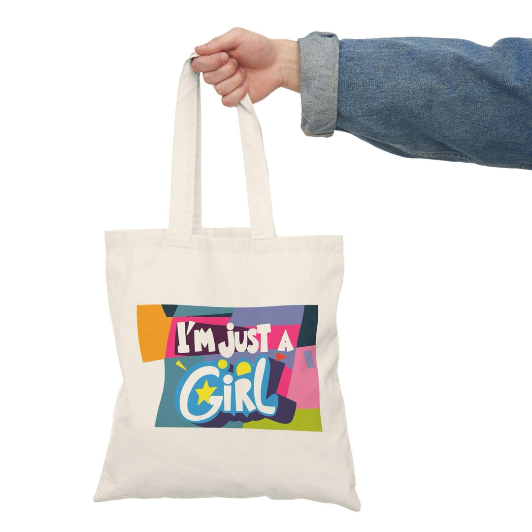 A cream-colored tote bag featuring a vibrant, multicolored text graphic that reads 'I’m Just a Girl.' The bright colors and playful font reflect a fun and youthful spirit, making it an excellent choice for everyday use