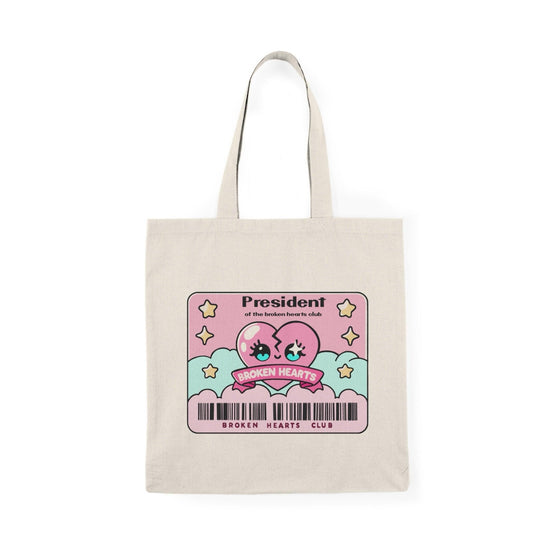 A cream-colored tote bag featuring a playful design that reads 'President of the Broken Hearts Club' surrounded by heart graphics. The whimsical design resonates with those who appreciate humorous and relatable messages about love and heartbreak