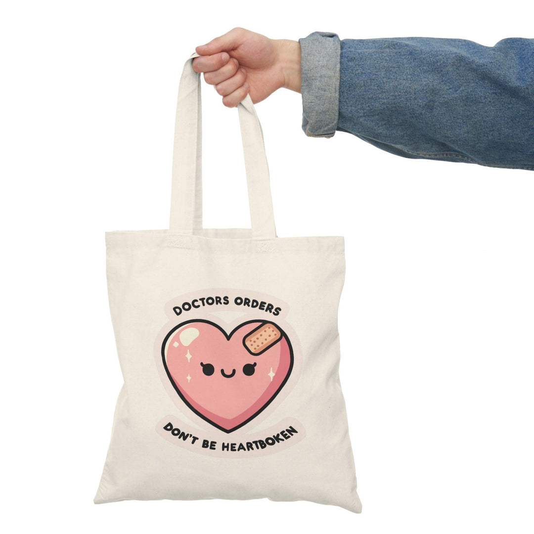 Tote bag featuring a playful and humorous design with the phrase 'Doctor's Orders: Don't Be Heartbroken.' The vibrant typography and fun graphics create a lighthearted accessory, perfect for adding a touch of humor and positivity to your everyday style while conveying a message of resilience."