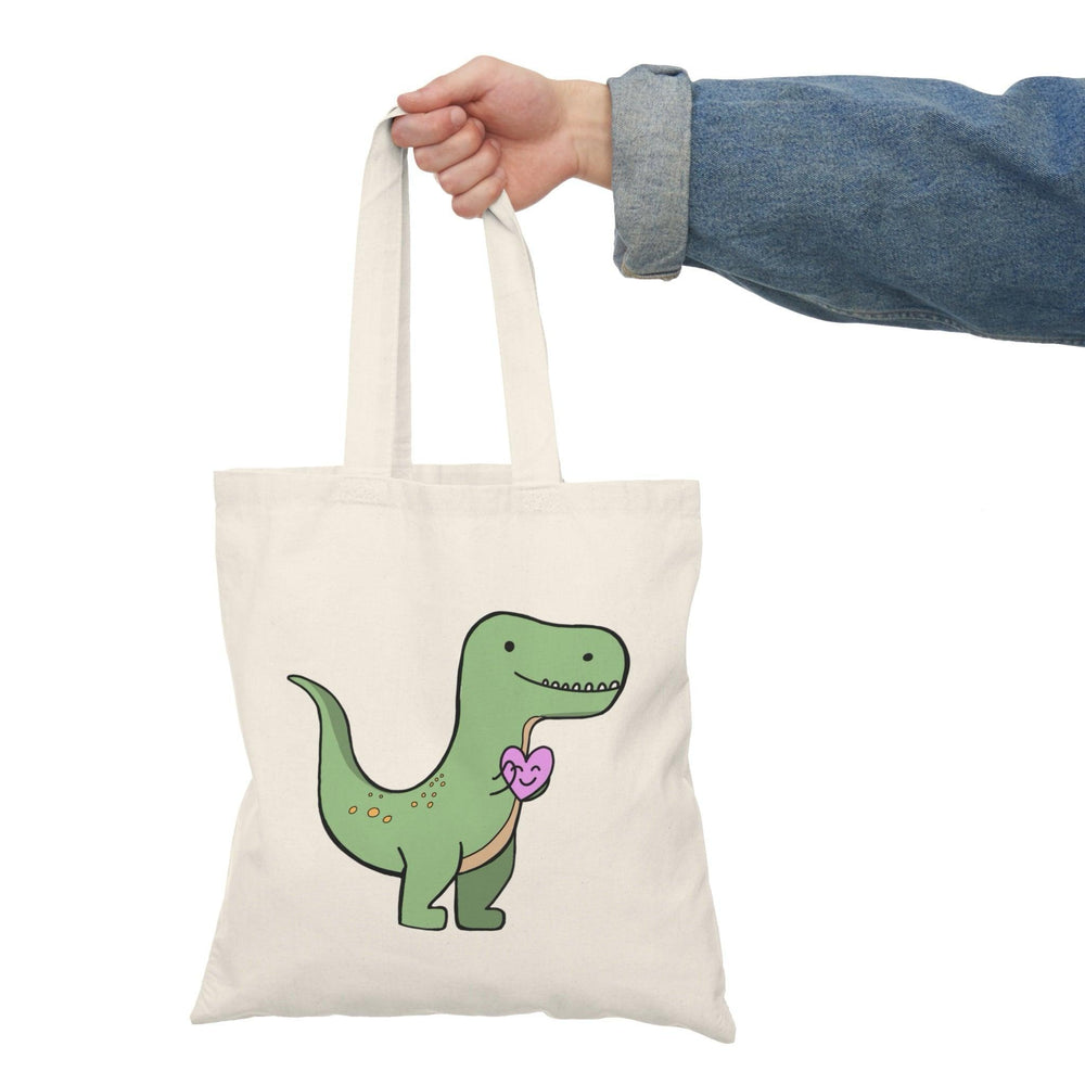 A natural canvas tote bag showcasing a charming graphic of a T-rex holding a pink heart. The cute and humorous design combines love and prehistoric fun, making it an ideal accessory for dinosaur lovers and those with a quirky sense of humor.