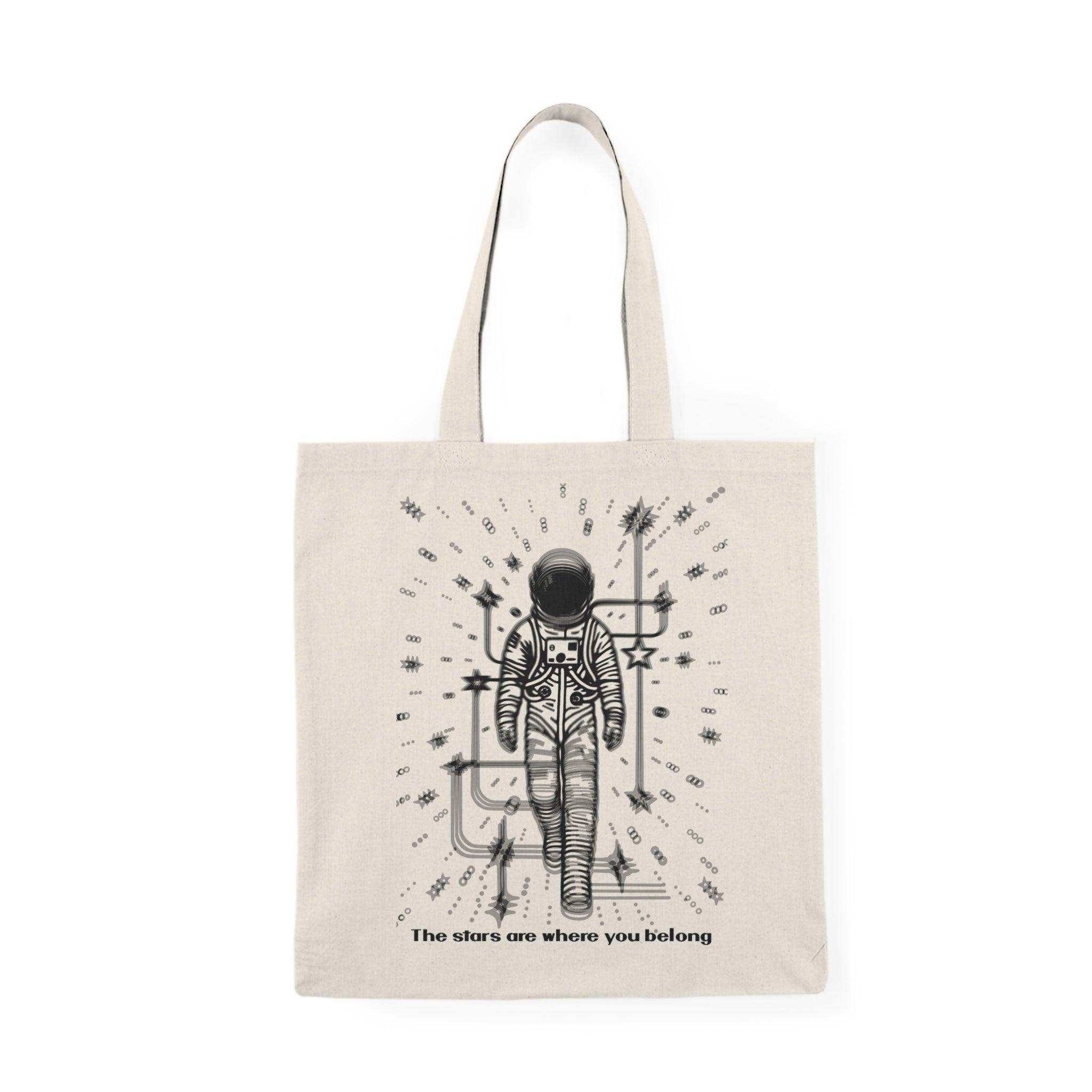 A cream-colored tote bag featuring a whimsical illustration of a person standing under a starry sky, with the phrase 'The stars are where you belong.' This poetic design captures a sense of wonder and belonging, making it perfect for dreamers and stargazers.