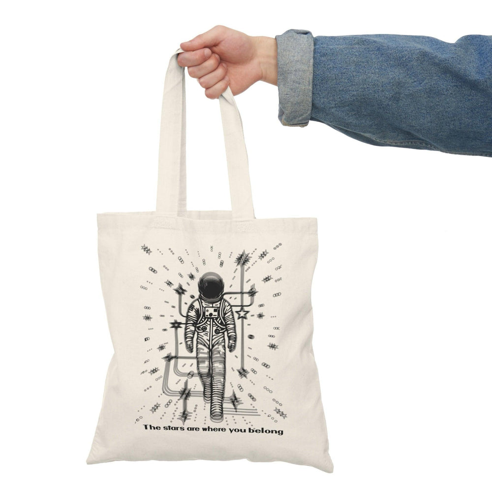 A cream-colored tote bag featuring a whimsical illustration of a person standing under a starry sky, with the phrase 'The stars are where you belong.' This poetic design captures a sense of wonder and belonging, making it perfect for dreamers and stargazers.