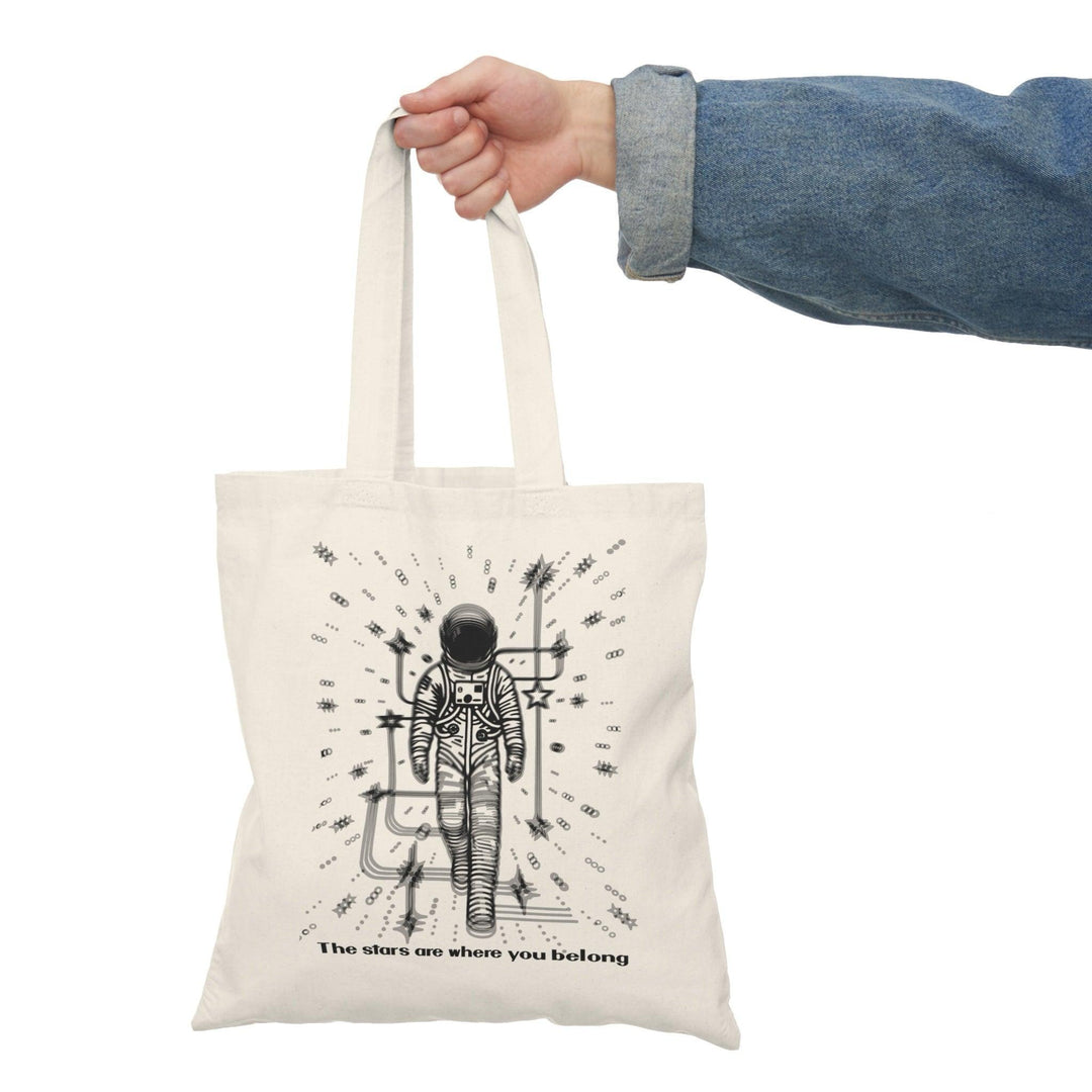 A cream-colored tote bag featuring a whimsical illustration of a person standing under a starry sky, with the phrase 'The stars are where you belong.' This poetic design captures a sense of wonder and belonging, making it perfect for dreamers and stargazers.