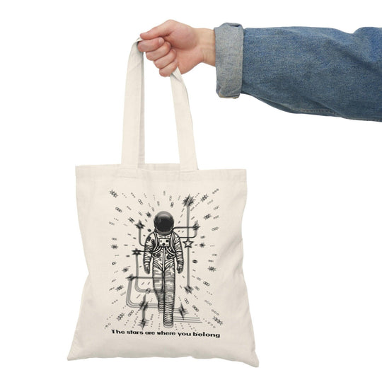 A cream-colored tote bag featuring a whimsical illustration of a person standing under a starry sky, with the phrase 'The stars are where you belong.' This poetic design captures a sense of wonder and belonging, making it perfect for dreamers and stargazers.