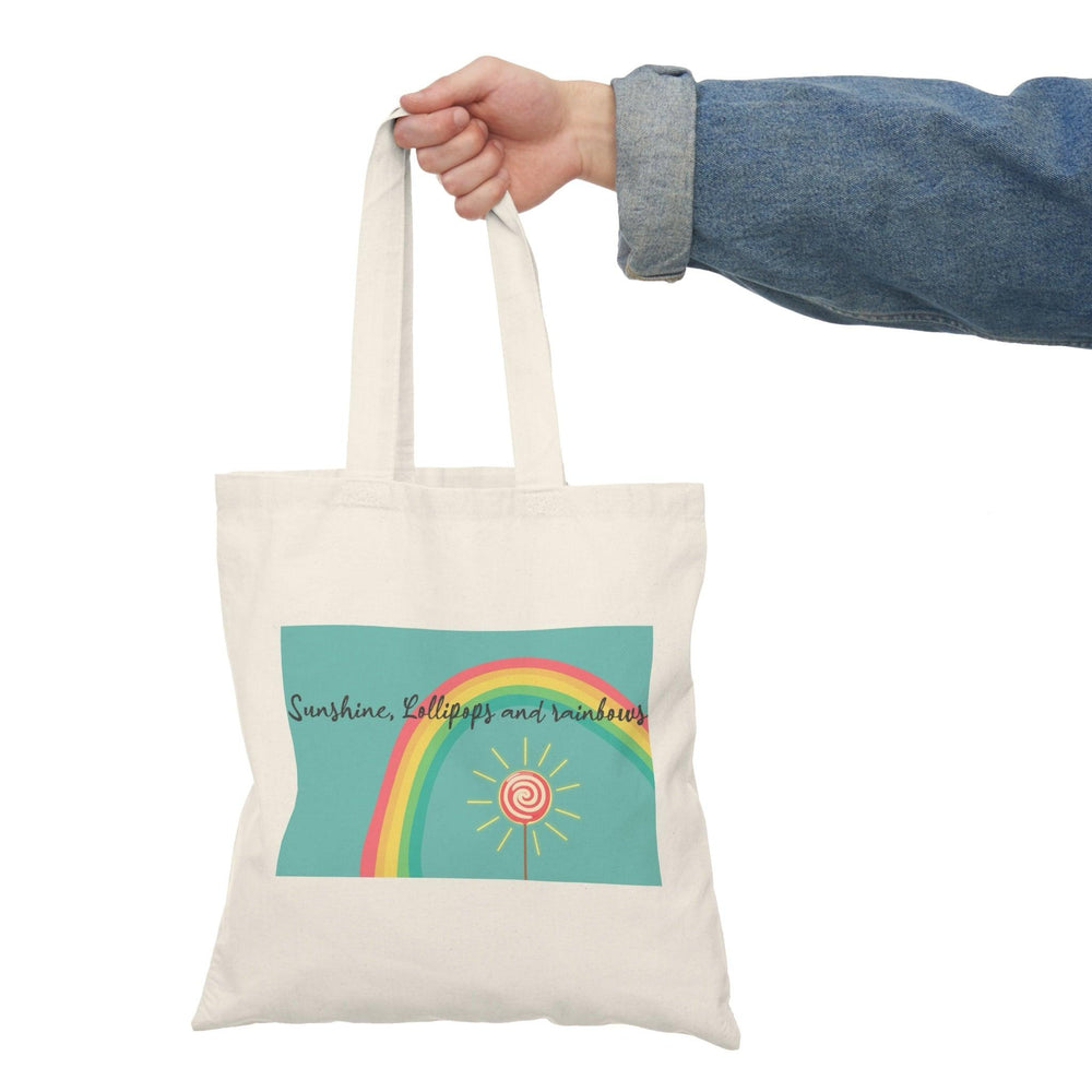 A cream tote bag adorned with a colorful graphic featuring a rainbow, lollipops, and sunshine with the text 'Sunshine, Lollipops and Rainbows.' This cheerful design radiates positivity and happiness, making it a delightful accessory for brightening up any day."