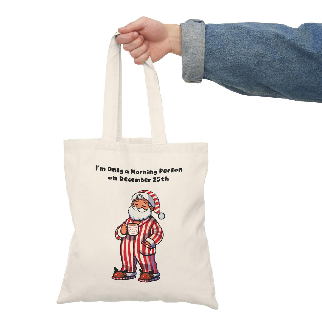 Funny Christmas tote bag with cozy Santa in striped pajamas and the phrase 'I'm Only a Morning Person on December 25th.' Perfect for holiday shopping, made from 100% recycled cotton