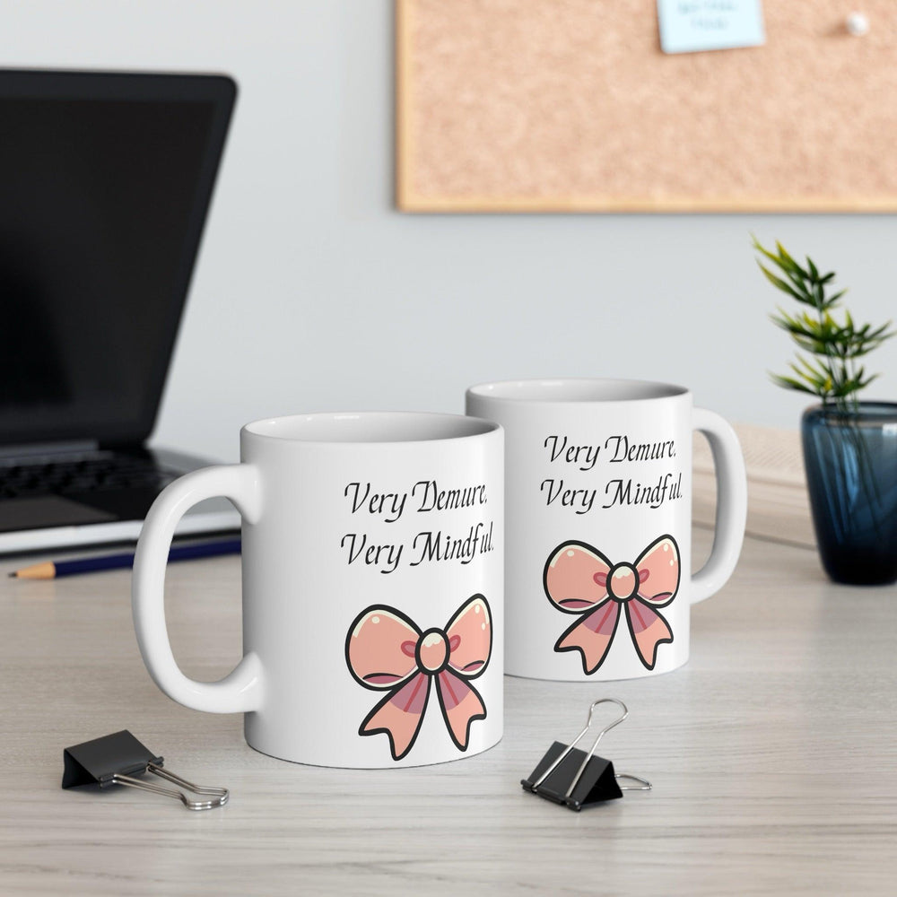White ceramic mug featuring a pair of pink bows and the phrase 'Very Demure, Very Mindful,' offering a subtle and stylish design for tea or coffee.