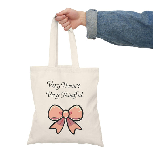 A cream tote bag adorned with a stylish design featuring the phrase 'Very Demure. Very Mindful.' paired with a decorative bow graphic. This elegant design resonates with those who appreciate a sense of calm and mindfulness in their lives