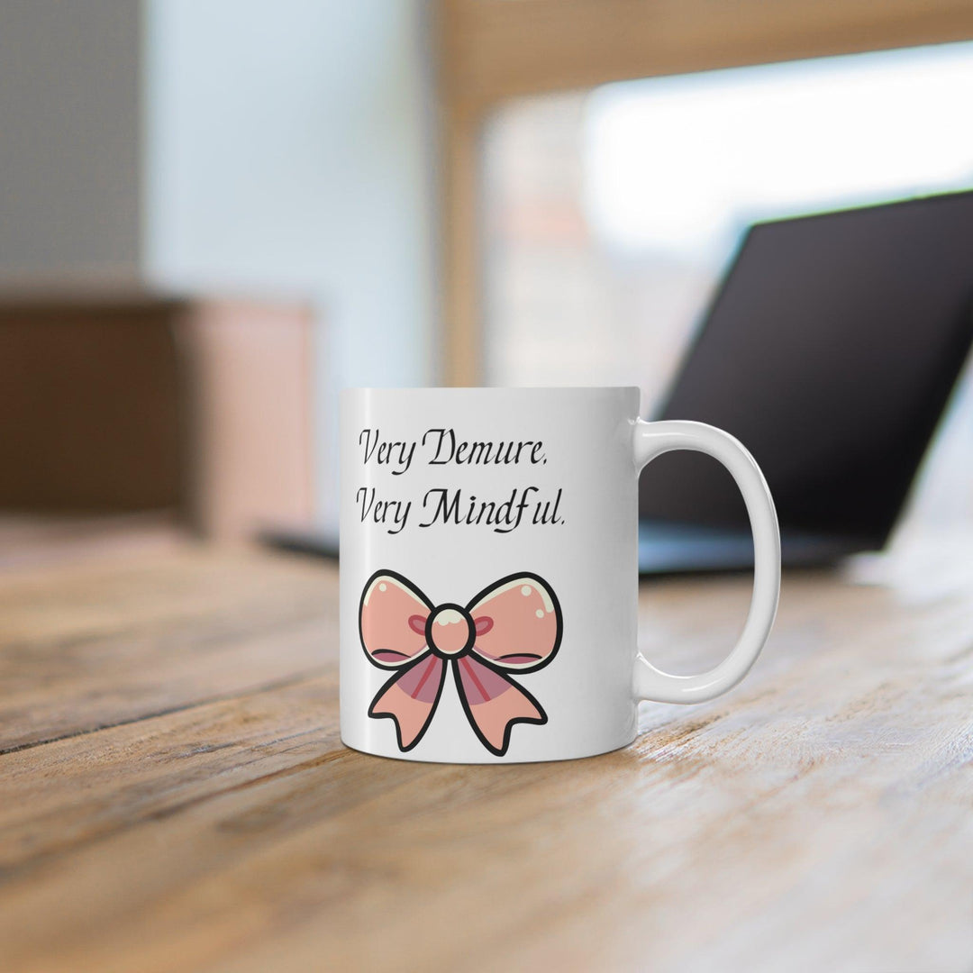 White ceramic mug featuring a pair of pink bows and the phrase 'Very Demure, Very Mindful,' offering a subtle and stylish design for tea or coffee.