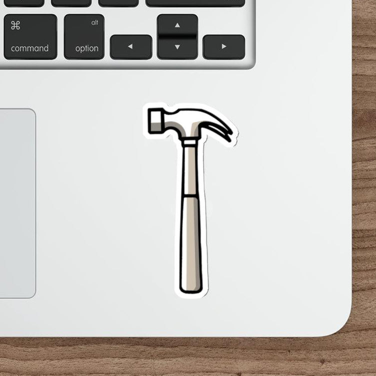 A waterproof sticker depicting a hammer, placed on a laptop next to a pen and a computer mouse, with a wooden texture background.