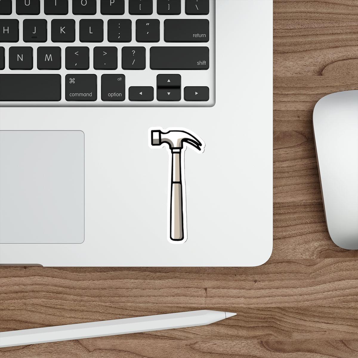 A waterproof sticker depicting a hammer, placed on a laptop next to a pen and a computer mouse, with a wooden texture background.