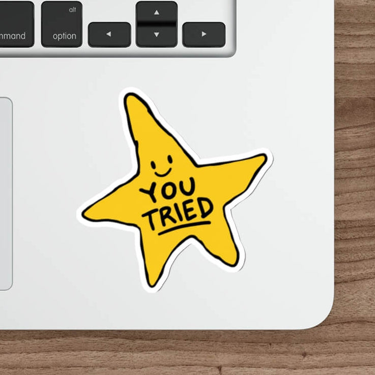 A yellow star-shaped sticker with a smiley face and the words 'YOU TRIED' in black, placed on the lower part of a silver laptop on a wooden desk next to a white mouse and stylus.