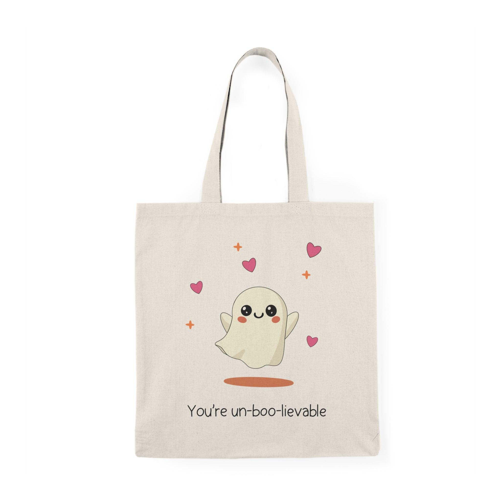 A natural tote bag adorned with an adorable illustration of a ghost surrounded by hearts and the text 'You're Un-boo-lievable.' This cute design captures a playful Halloween spirit, making it a fun accessory for the spooky season or any ghost lover