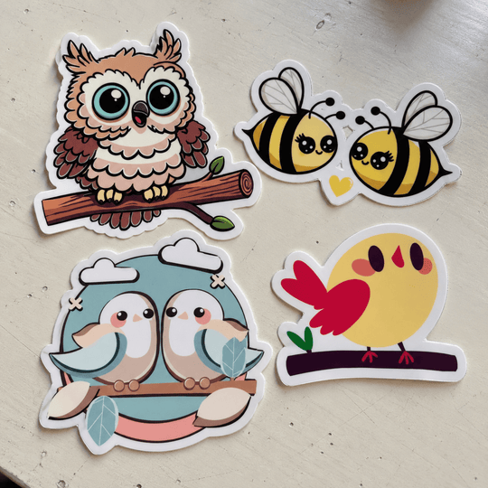 Four cartoon animal stickers arranged on a light beige surface: an owl on a branch, two bees facing each other, two birds hugging, and a yellow bird with a red tail. The stickers have white borders and are photographed from above.