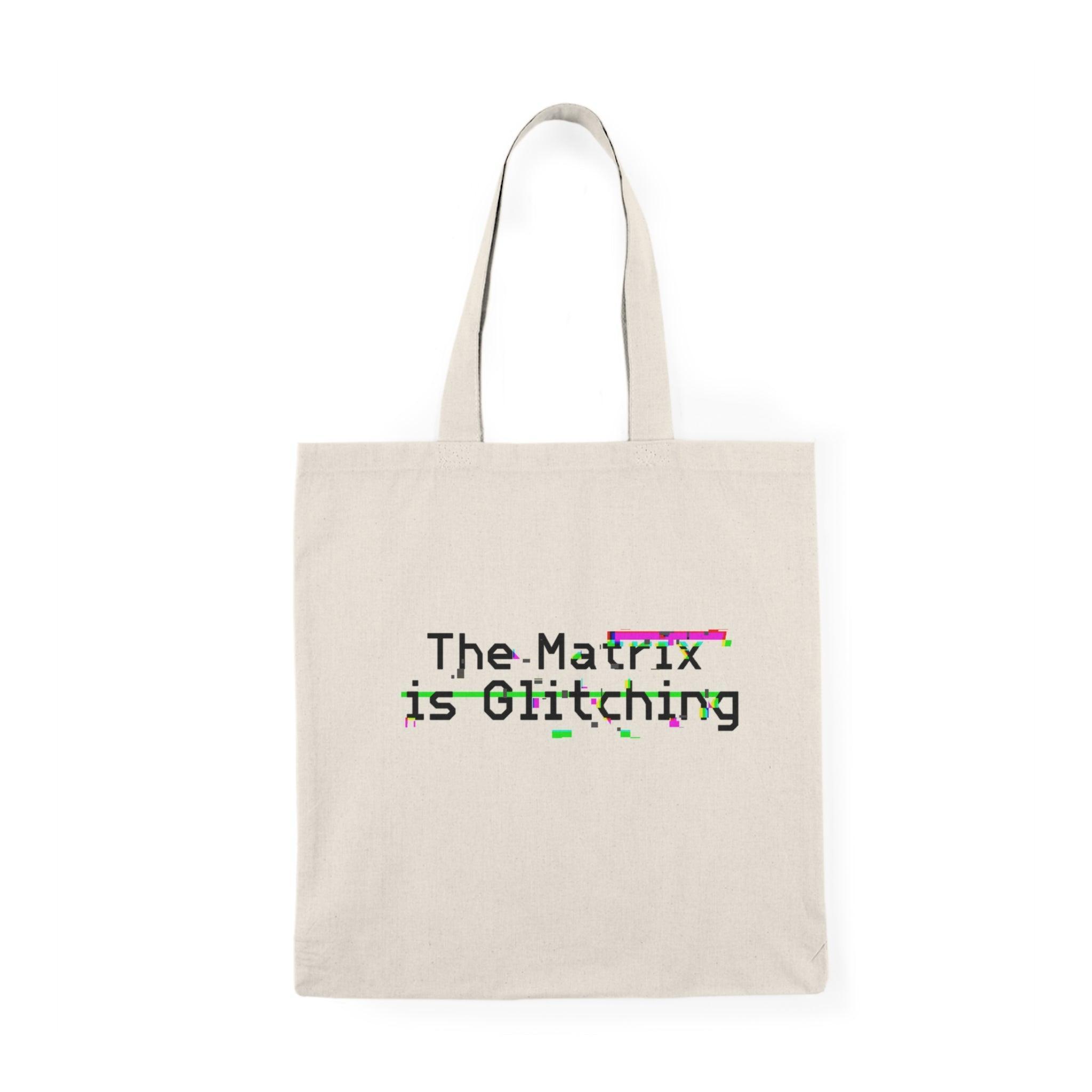 A cream-colored tote bag featuring the phrase 'The Matrix is Glitching' in a dynamic glitch graphic, capturing the essence of digital anomalies. The design uses an array of weird colors to embody the unsettling yet fascinating concept of reality distortion. The tote measures 40 x 38 cm (15.7 x 15 inches) and is made from 100% recycled cotton, providing an eco-friendly choice without sacrificing durability or style.