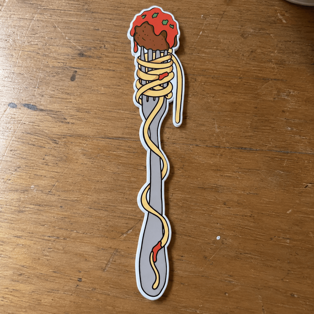 A die-cut sticker shaped like a silver fork wrapped in spaghetti with a meatball and tomato sauce topping, placed on a wooden tabletop.
