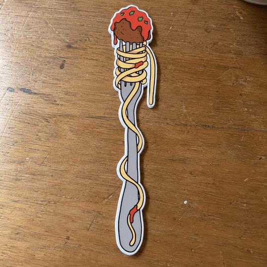 A die-cut sticker shaped like a silver fork wrapped in spaghetti with a meatball and tomato sauce topping, placed on a wooden tabletop.
