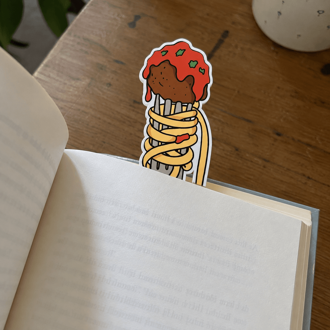 A die-cut sticker shaped like a spaghetti cone with red sauce and green herbs, placed on an open book on a wooden table.