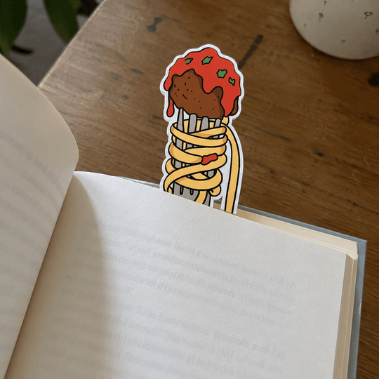 A die-cut sticker shaped like a spaghetti cone with red sauce and green herbs, placed on an open book on a wooden table.