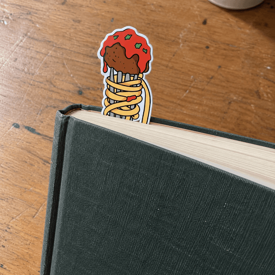 A die-cut sticker featuring an illustrated spaghetti and meatball design, clipped onto the edge of a dark green hardcover book on a wooden tabletop.