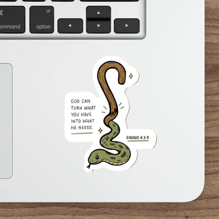 A die-cut sticker featuring a cartoon green and brown snake with the text 'GOD CAN TURN WHAT YOU DO INTO WHAT HE NEEDS' and 'EXODUS 4:2-4' on a white laptop palm rest, shown on a wooden desk next to a keyboard, mouse, and stylus.