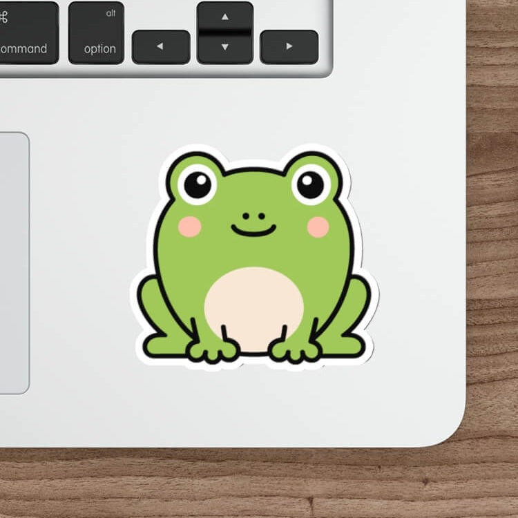 A cute green cartoon frog sticker with a white belly and black outline, applied to the palm rest of a silver laptop. The laptop is shown on a wooden desk alongside a white mouse, a stylus, and part of a keyboard.