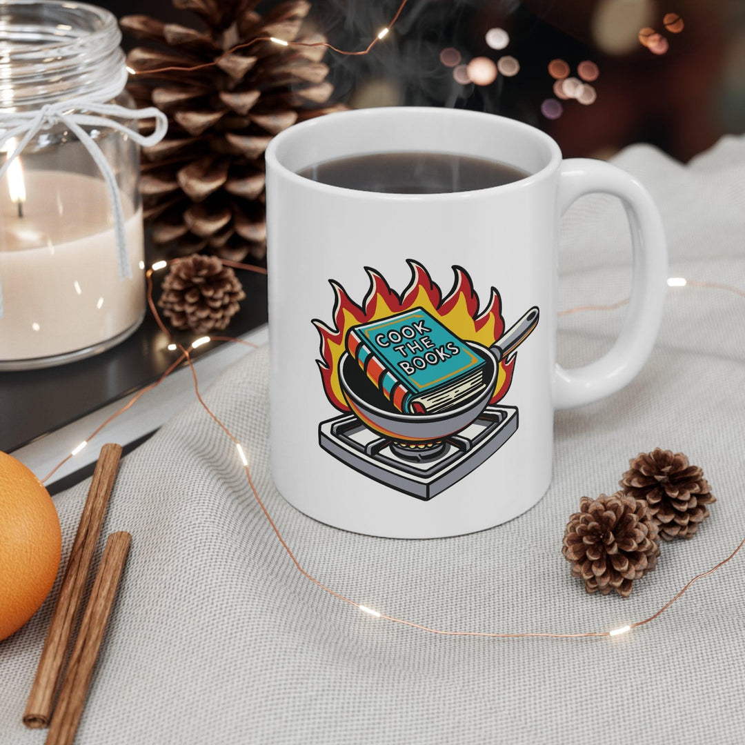 Quirky white ceramic mug featuring a cartoon book engulfed in flames, with the clever text 'Cook the Books,' making it a fun gift for book lovers