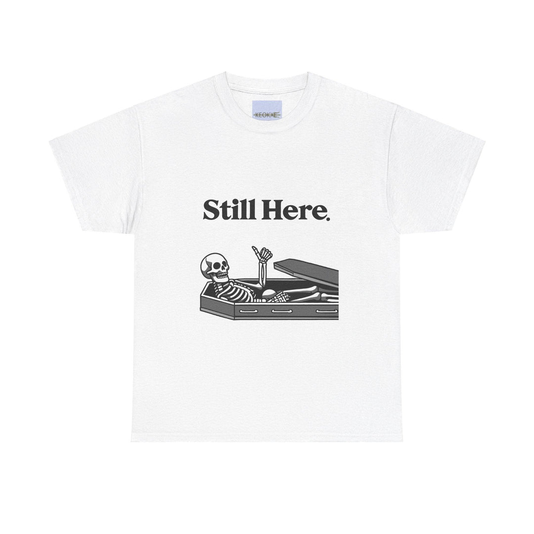 White T-shirt featuring a laid-back skeleton lounging on a chaise with the phrase 'Still Here.' A light-hearted design, perfect for embracing perseverance with a twist of humor."