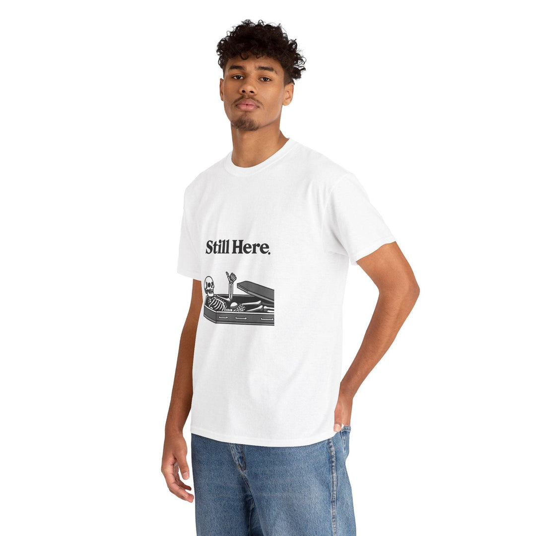 White T-shirt featuring a laid-back skeleton lounging on a chaise with the phrase 'Still Here.' A light-hearted design, perfect for embracing perseverance with a twist of humor."