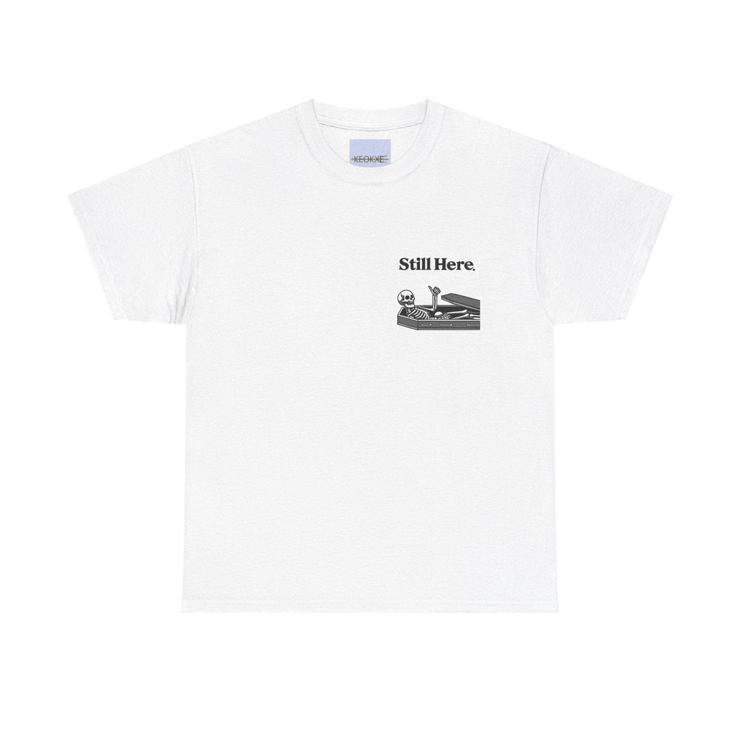 White T-shirt featuring a laid-back skeleton lounging on a chaise with the phrase 'Still Here.' A light-hearted design, perfect for embracing perseverance with a twist of humor."