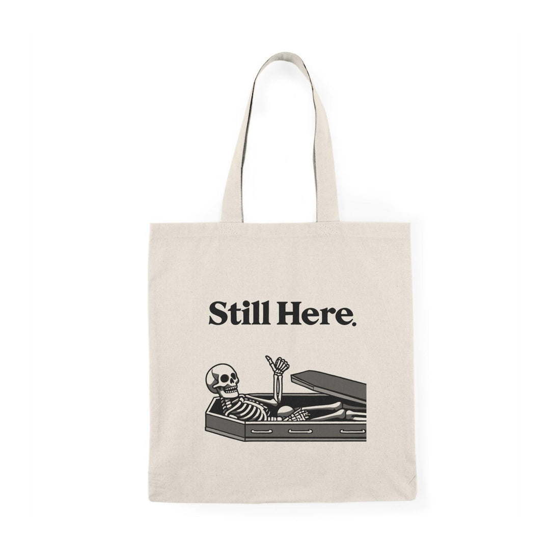 A natural canvas tote bag featuring an illustration of a skeleton lying in a coffin with the text 'Still Here.' The humorous design adds a touch of gothic charm, making it a fun accessory for those who enjoy Halloween-themed or quirky designs