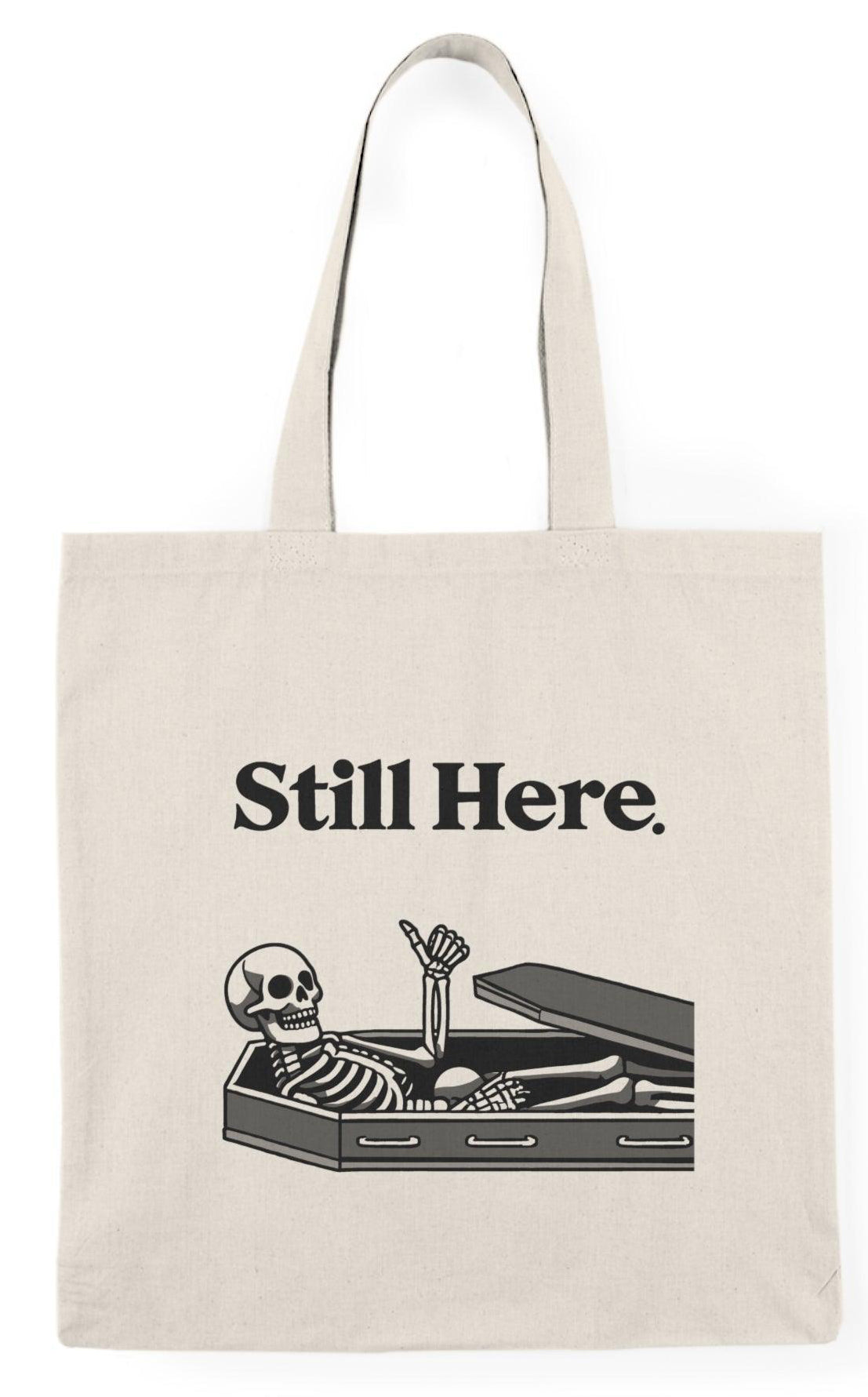 A natural canvas tote bag featuring an illustration of a skeleton lying in a coffin with the text 'Still Here.' The humorous design adds a touch of gothic charm, making it a fun accessory for those who enjoy Halloween-themed or quirky designs KEOLIX 