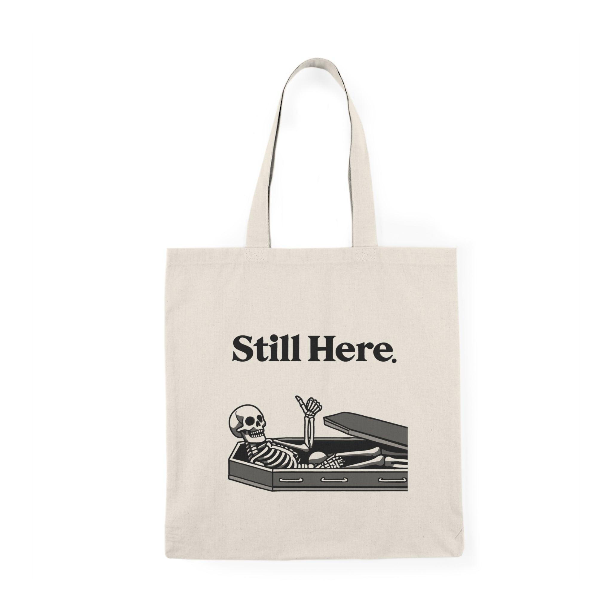 A natural canvas tote bag featuring an illustration of a skeleton lying in a coffin with the text 'Still Here.' The humorous design adds a touch of gothic charm, making it a fun accessory for those who enjoy Halloween-themed or quirky designs