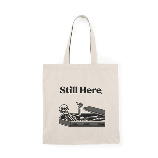 A natural canvas tote bag featuring an illustration of a skeleton lying in a coffin with the text 'Still Here.' The humorous design adds a touch of gothic charm, making it a fun accessory for those who enjoy Halloween-themed or quirky designs